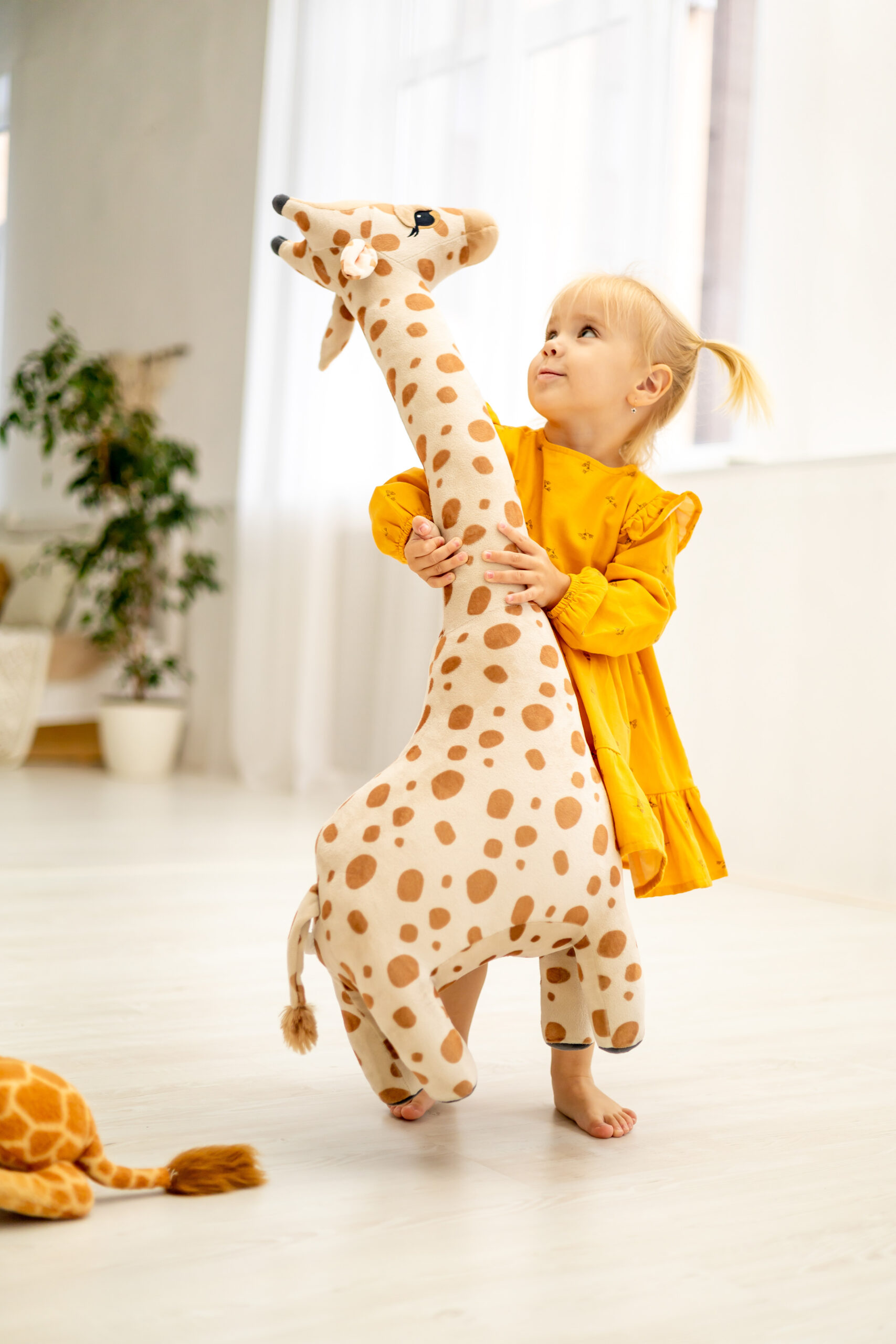 a little child girl in a yellow dress plays with a giraffe at home measuring her height in a bright room and smiling, happy childhood